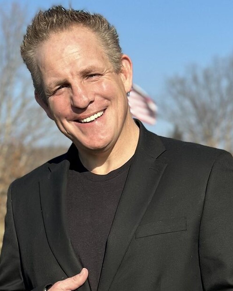 Grant Whitmer, smiling, in a dark blazer outdoors on a sunny winter day