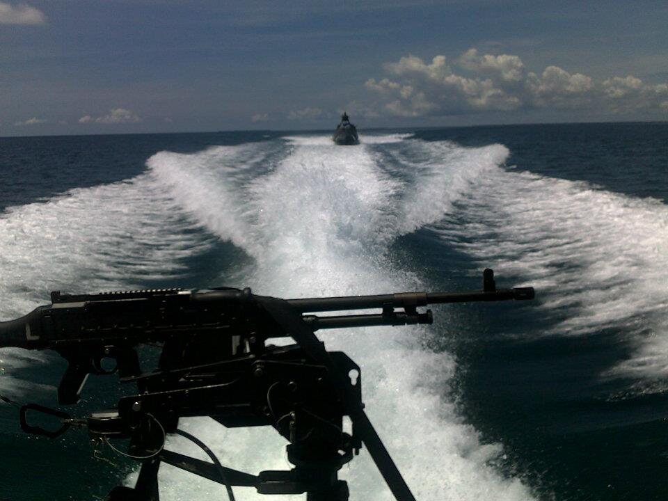 Looking back over a boat's mounted gun at its wake, a second boat following in the distance